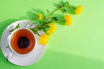 A Cup of black tea and yellow flowers of Trollius europaeus on a green background. Evening and morning tea. Copy space.