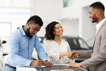 Spouses Shaking Hands With Auto Seller Signing Papers In Dealership