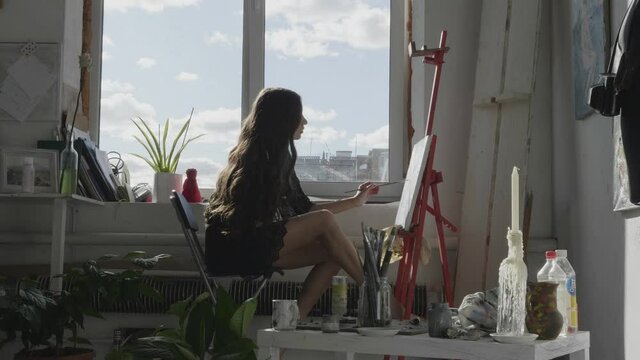 attractive brunette woman in short dress works on picture with brush sitting near large window on sunny day slow motion