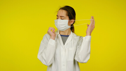 Young female infectious disease doctor removes a protective medical mask from her face. Nurse in a white coat on a yellow background after the patient