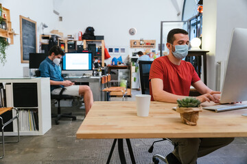 Millennials men with surgical mask at working at computer in office at distance for protection and...