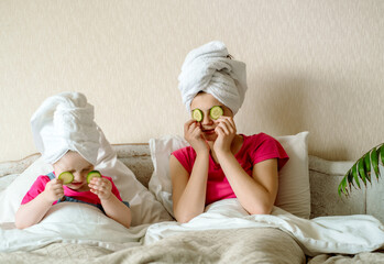 Obraz na płótnie Canvas Morning skin care. two girls in bed make a mask of cucumber. white towel on hair after shower, bright room. first makeup for a teenage girl. game for baby.