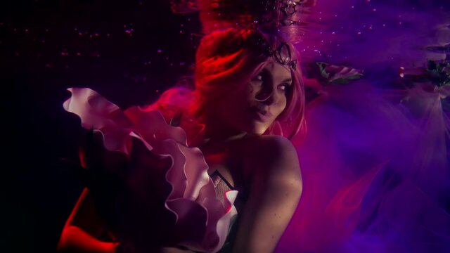Portrait in profile of a beautiful girl who is under water on a dark background, she has a tiara on her head, around her dress fabric, floating flower buds. She is holding a huge Bud in her hands.