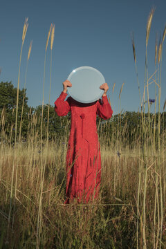 Teenage girl in red dress covering her face with mirror while standing in field