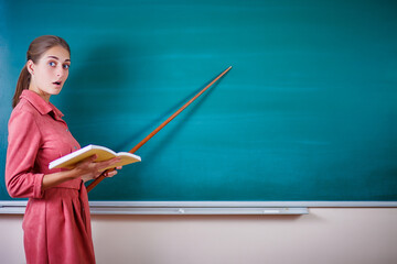 Young woman teacher stands at a blackboard with a pointer. Template for the school timetable and other information. Beautiful tacher shows on empty copy space on the school board. Education concept.