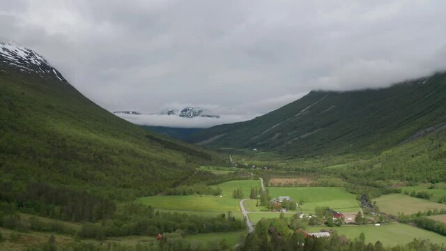 Lush, green Innerdalen valley surrounded by mountains. A patchwork of farm fields and pastures. Heavy white clouds hanging above the valley. Snowcowered mountains towering on the horizon.