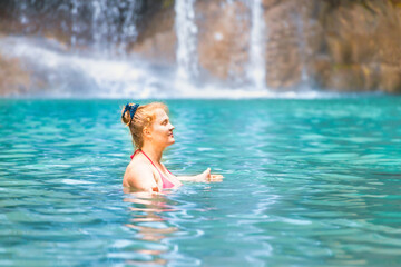 Red haired young woman in pink bikini swimsuit relaxes in emerald tropical lake with waterfall. Erawan National park, Kanchanaburi, Thailand