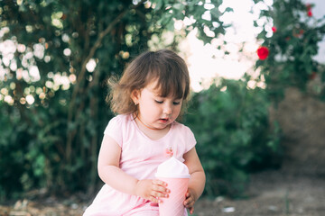 A beautiful little girl in a pink dress against a background of plants drinks a drink from a cocktail glass