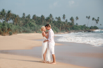 Fototapeta na wymiar Wedding concept. Young caucasian just married couple kissing on the beach, stand barefoot in wedding clothes nea the sea and palm trees. Beach wedding concept