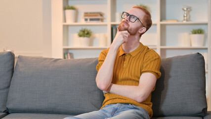 Pensive Redhead Man Sitting on Sofa at Home and Thinking 