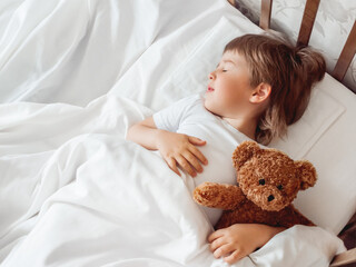 Toddler sleeps in bed with cute teddy bear. Little boy under white blanket with fluffy toy. Plush...