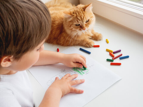 Left-handed toddler draws colorful robot. Kid uses wax crayons to paint it. Cute ginger cat lies on window sill with child. Coloring pages to train fine motor skills.