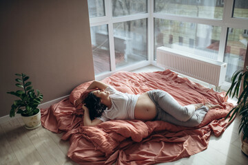 Pretty young brunette pregnant woman laying by the window in a big light room. She is touching a belly by hand. Sun comes through the window. Bed linen of lush lava colour. Relaxing at Home