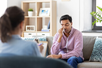 psychology, mental therapy and people concept - crying young indian man patient wiping tears with paper tissue and woman psychologist at psychotherapy session