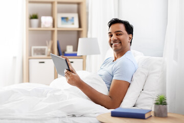 people, bedtime and rest concept - happy smiling indian man with tablet pc computer lying in bed at home