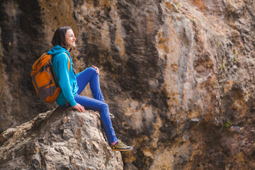 Naklejka na ściany i meble A woman with a backpack sits on a large stone on a background of rocky mountains.