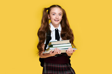 Fototapeta premium Excited happy elementary schoolgirl holding books on vivid yellow background, pupil wearing uniform with school stuff
