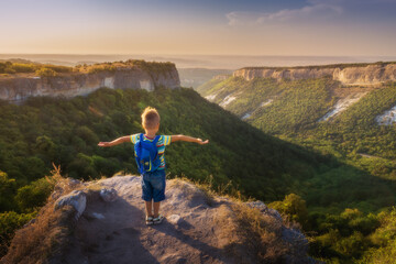 Cute little boy of 4 years old with a backpack stands on the edge of a cliff, arms outstretched, and looks at a stunning panorama of mountains at sunset. Hiking and travelling with children 