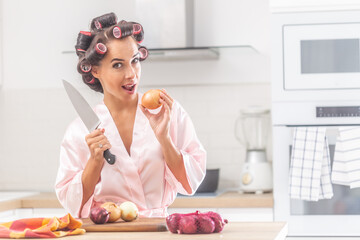 Good looking woman stands in the kitchen holding knife and an onion, wearing hair curlers and night...