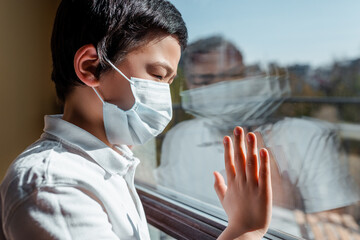 upset asian child in medical mask looking through window on quarantine
