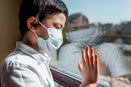 Upset Asian Child In Medical Mask Looking Through Window On Quarantine