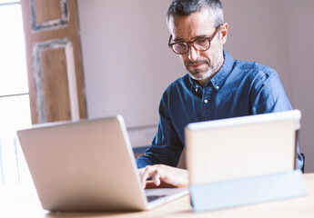 Good looking middle aged modern man having an online meeting on his laptop and tablet from home with his colleagues