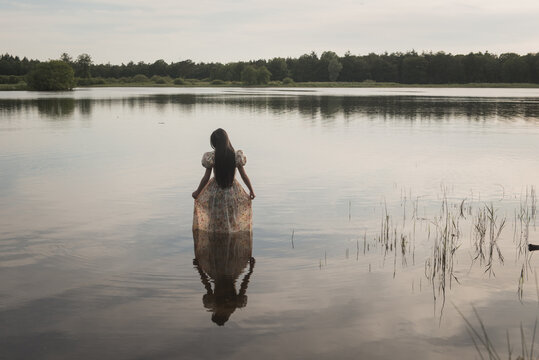 Girl with long hair wading in water wearing a classic romantic dress during sunset