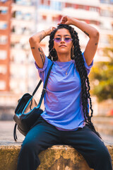 Lifestyle, look of young dark-skinned woman with long braids and purple glasses sitting on a bench in the park, with buildings in the background