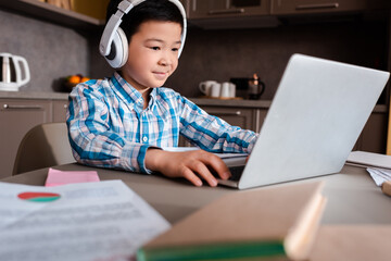 smiling asian boy studying online with books, laptop and headphones at home during quarantine