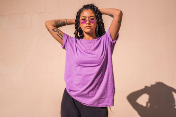 Urban session. portrait of young dark-skinned woman with long braids wearing purple glasses on a plain cream-colored background, with seductive look