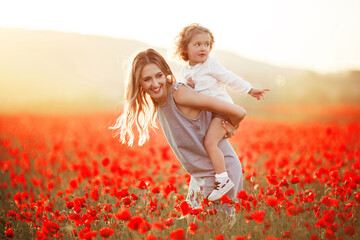 Beautiful smiling child girl with mother are having fun in field of red poppy flowers over sunset...