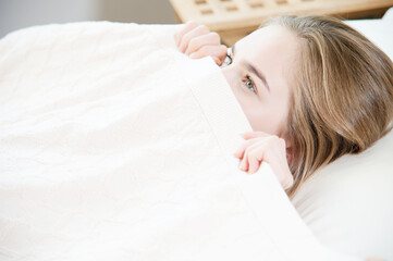 A young attractive woman peeks out from under the blankets hiding behind them with fear when he wakes up. The concept of female fears related to sleep and intimate life in the family