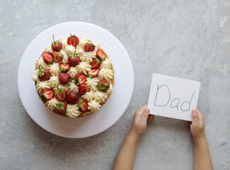 In the hands of the child a card with the inscription dad. A gift for dad cake with fresh strawberries. Image with copy space, top view, horizontal orientation.