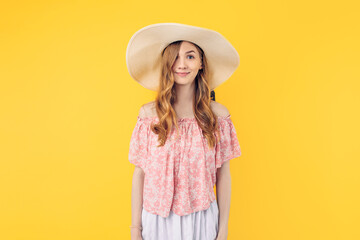 A happy attractive young woman in a summer dress and hat poses standing on an yellow background