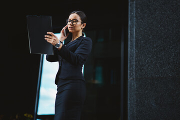 Busy female entrepreneur dressed in stylish elegant clothing reading business information from textbook and discussing it with partners via modern smartphone device.Publicity area for advertising