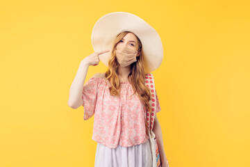 Happy young woman in a summer hat, with a protective medical mask on her face, on an isolated yellow background