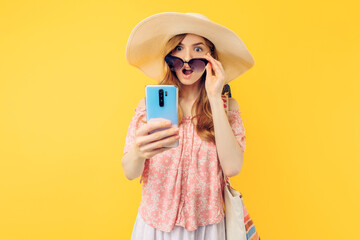 A shocked surprised woman in a summer hat and sunglasses with a mobile phone on an yellow background