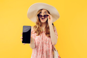 An excited shocked woman in a summer hat and sunglasses shows a blank mobile phone screen for a copy of the space on an yellow background