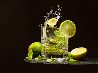 Glass of Mojito cocktail with fresh limes,mint leaves,ice cubes and refreshing water.Splash of cocktail Mojito on black stone plate.