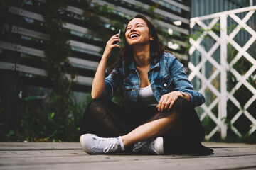 Happy young woman with gorgeous long hair laughing during mobile conversation via smartphone device sitting in lotus position.Cheerful female with mobile phone in hand having fun in free time outdoors