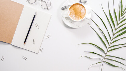 Flat lay white minimalistic office work space desk with copy space background. Work place with cappuccino cup coffee, notebook, glasses and palm branch.