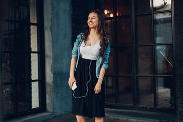 Happy attractive hipster girl in denim jacket and black skirt listening audio songs in modern earphones connected to mobile phone.Female meloman enjoying music in headphones in leisure time