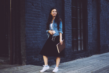 Stylish smiling student with textbook in hand listening audio in earphones connecting to mobile phone while strolling in street in urban setting and looking away.Promotional background for advertising