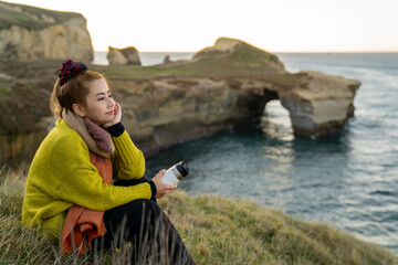 Beautiful asian woman with coffee cup during sunrise at Tunnel beach, Otago Peninsula, New Zealand.