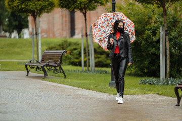 A masked girl is walking along the street. A girl in a protective mask walks in the park with an...