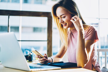 Positive female blogger reading news from social networks browsing webpage on smartphone device.Smiling hipster girl happy about good news getting during online chatting with friend on telephone