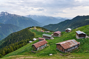 Landscape photo of Pokut Plateau with traditional wooden houses, snowy mountains, clouds and forest. Taken in summer at northeastern Black Sea / Karadeniz region of Turkey