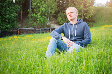 Naklejka na ściany i meble The concept of walking alone. Young caucasian blond man in jeans sits in a clearing against the background of the forest. A cozy image in calm warm colors.
