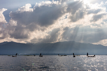 Fishermen on fishing boats made of teak wood and covered by lacquer. Traditional apparels: hat, fishing net and paddles. Cloudy sky, light filtered through clouds. Inle lake, Myanmar, southeast Asia