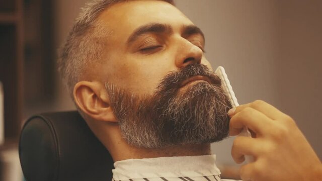 Barber shaving bearded man in barbershop. Hairdresser making fashionable beard with brush and razor. Male skin care and beard style concept. Close up of hipster male